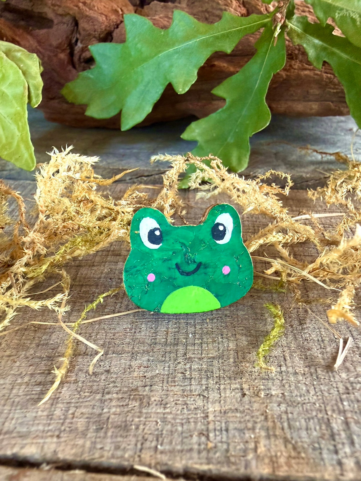 Handpainted pink cheeked frog cork pin badge face-on on weathered wood with dried moss and oak leaves — bright green frog with rosy pink cheeks