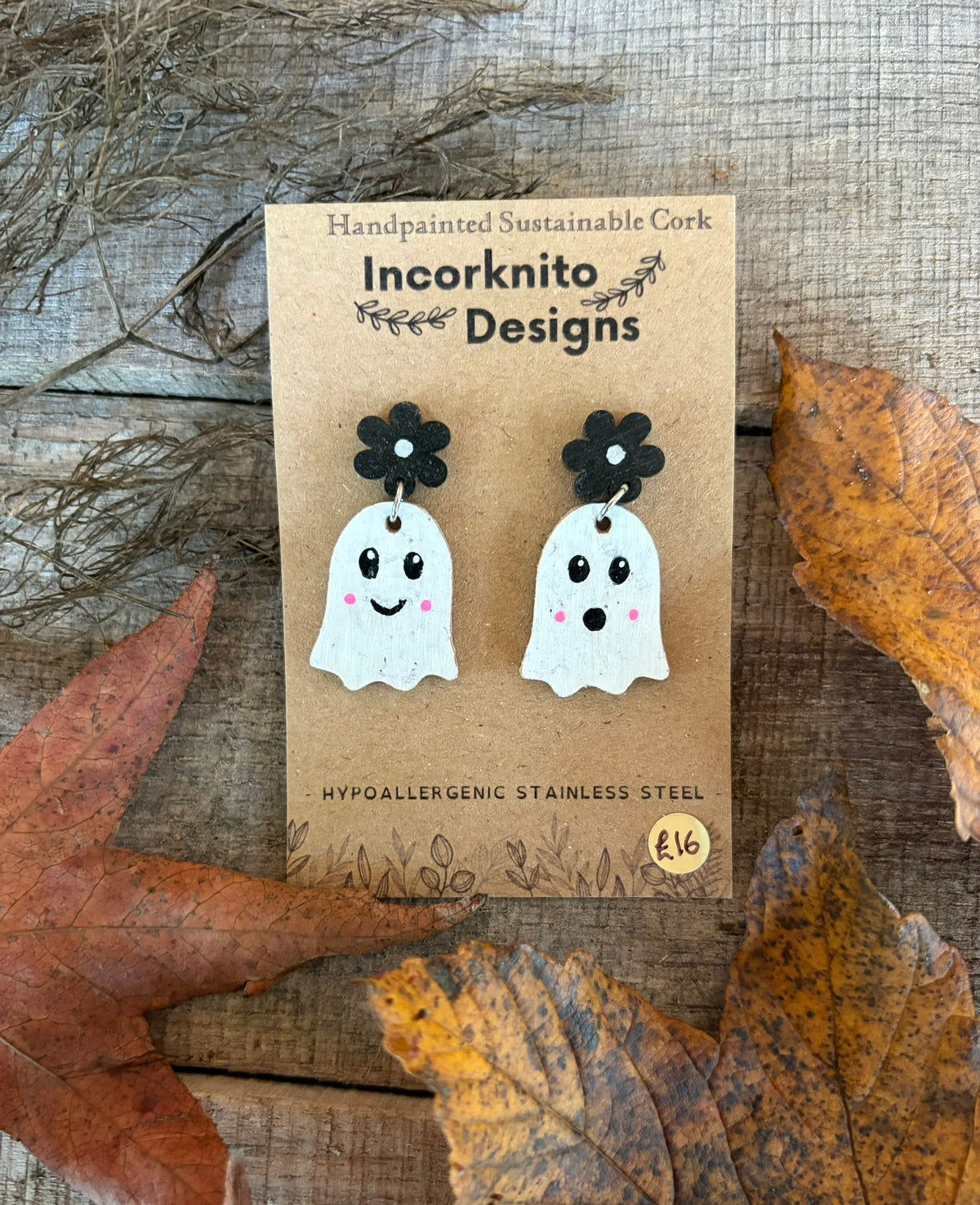 White ghost earrings with black flower stud tops and pink cheeks, displayed on a kraft card among autumn leaves.