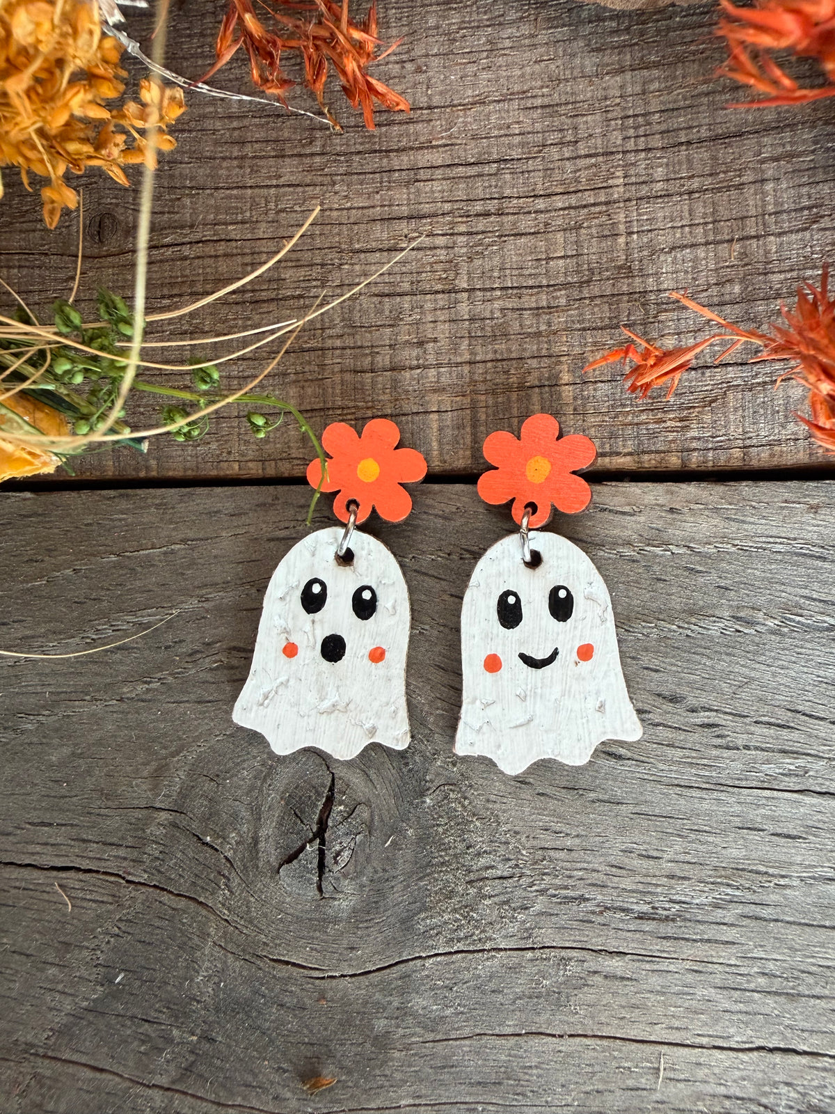 Close-up of orange flower ghost earrings on wood, showing one smiling face and one surprised face.