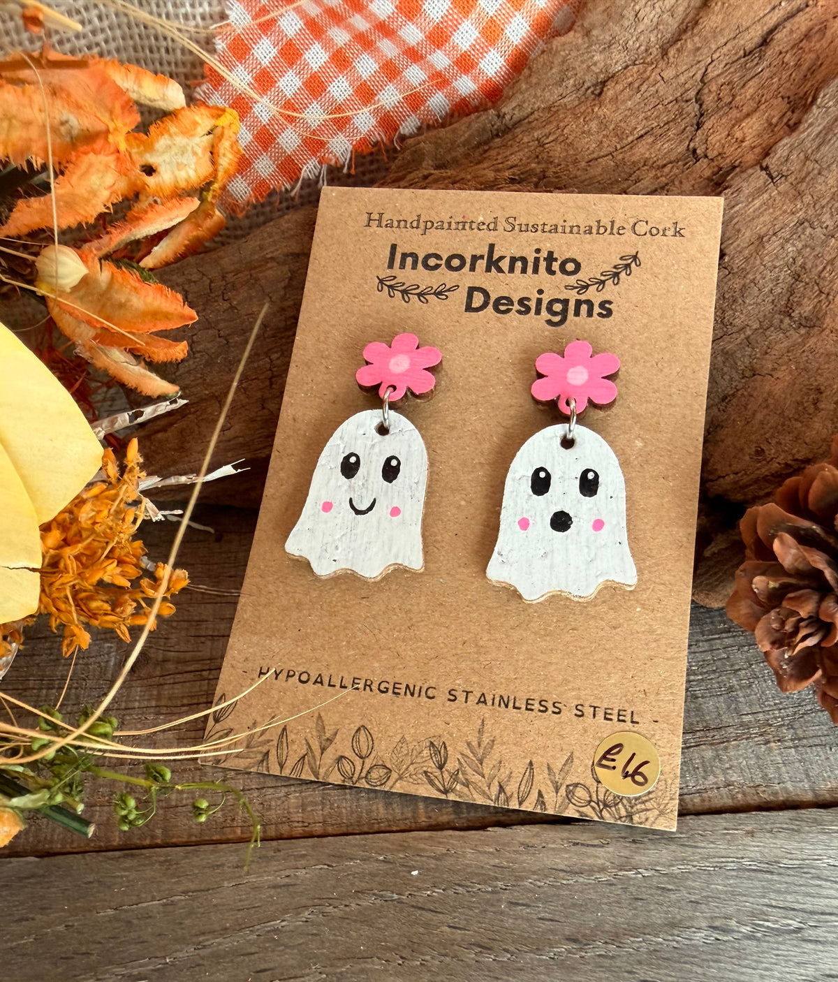 White handpainted cork ghosts with pink flower studs and pink cheeks, on a display card beside dried flowers.