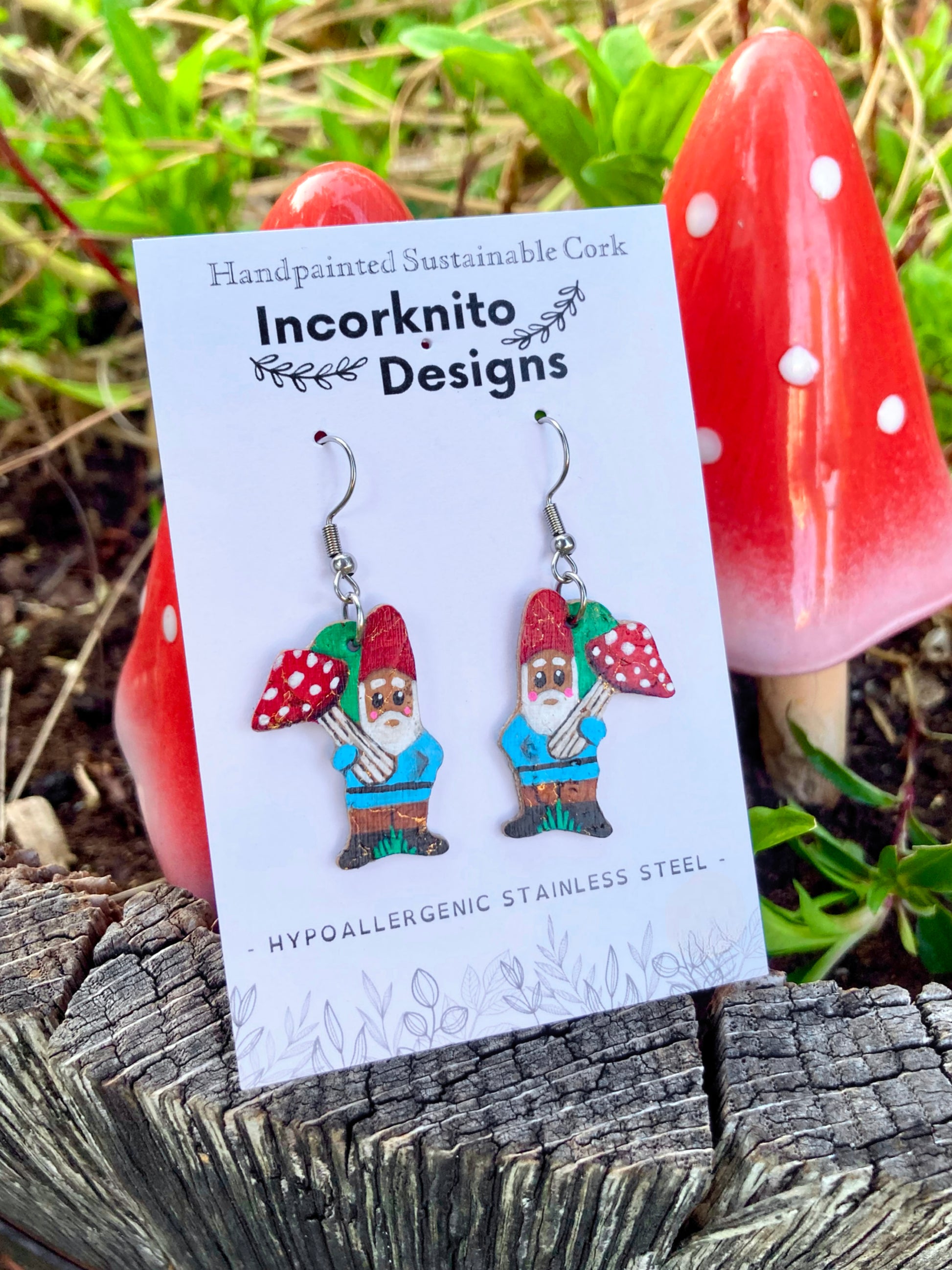 Handpainted gnome and toadstool cork earrings on silver hooks on Incorknito Designs card, surrounded by ceramic fly agaric toadstools on weathered wood