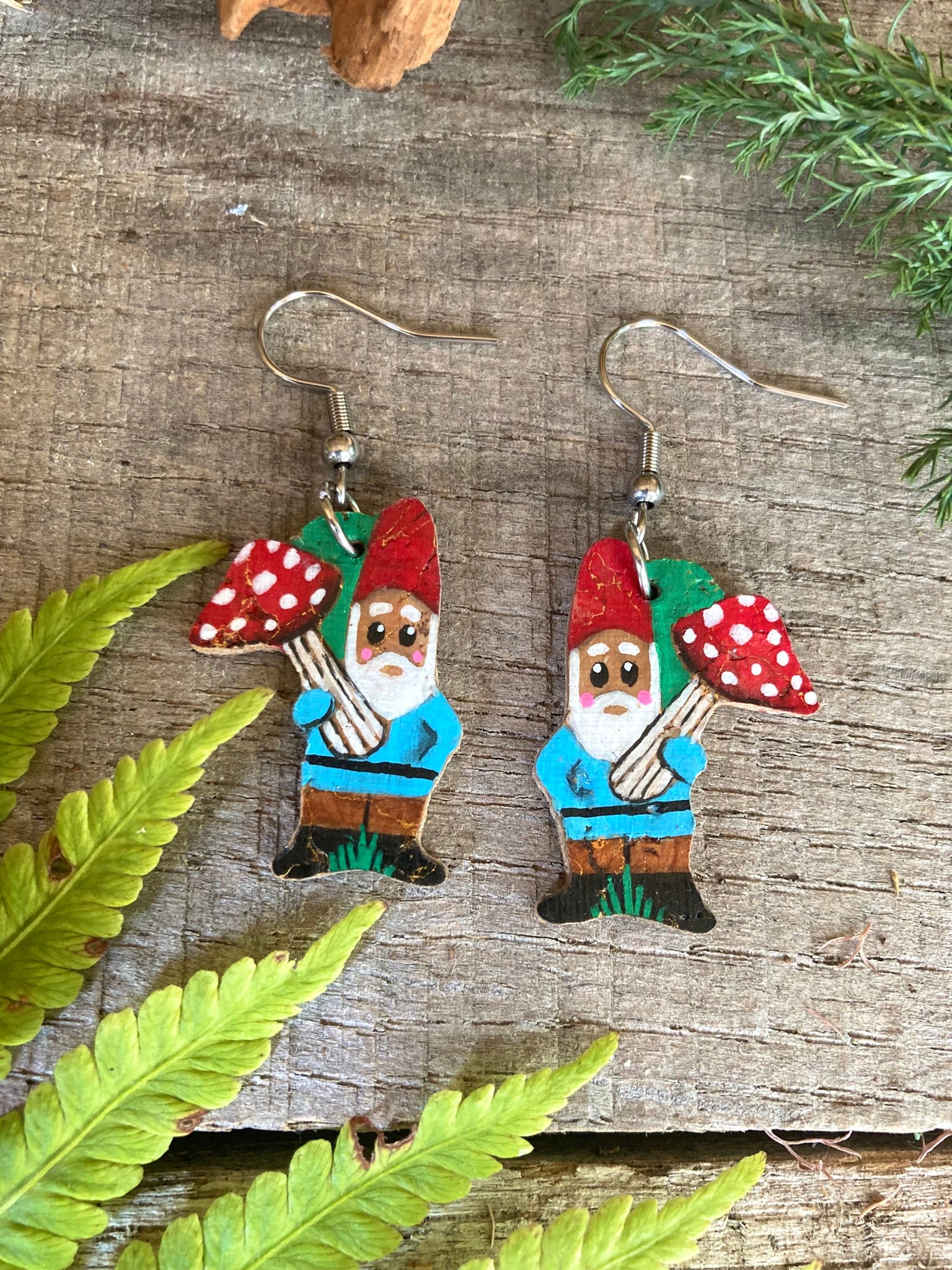 Flat lay of handpainted gnome and fly agaric toadstool cork earrings on silver hooks on grey weathered wood with fern leaves