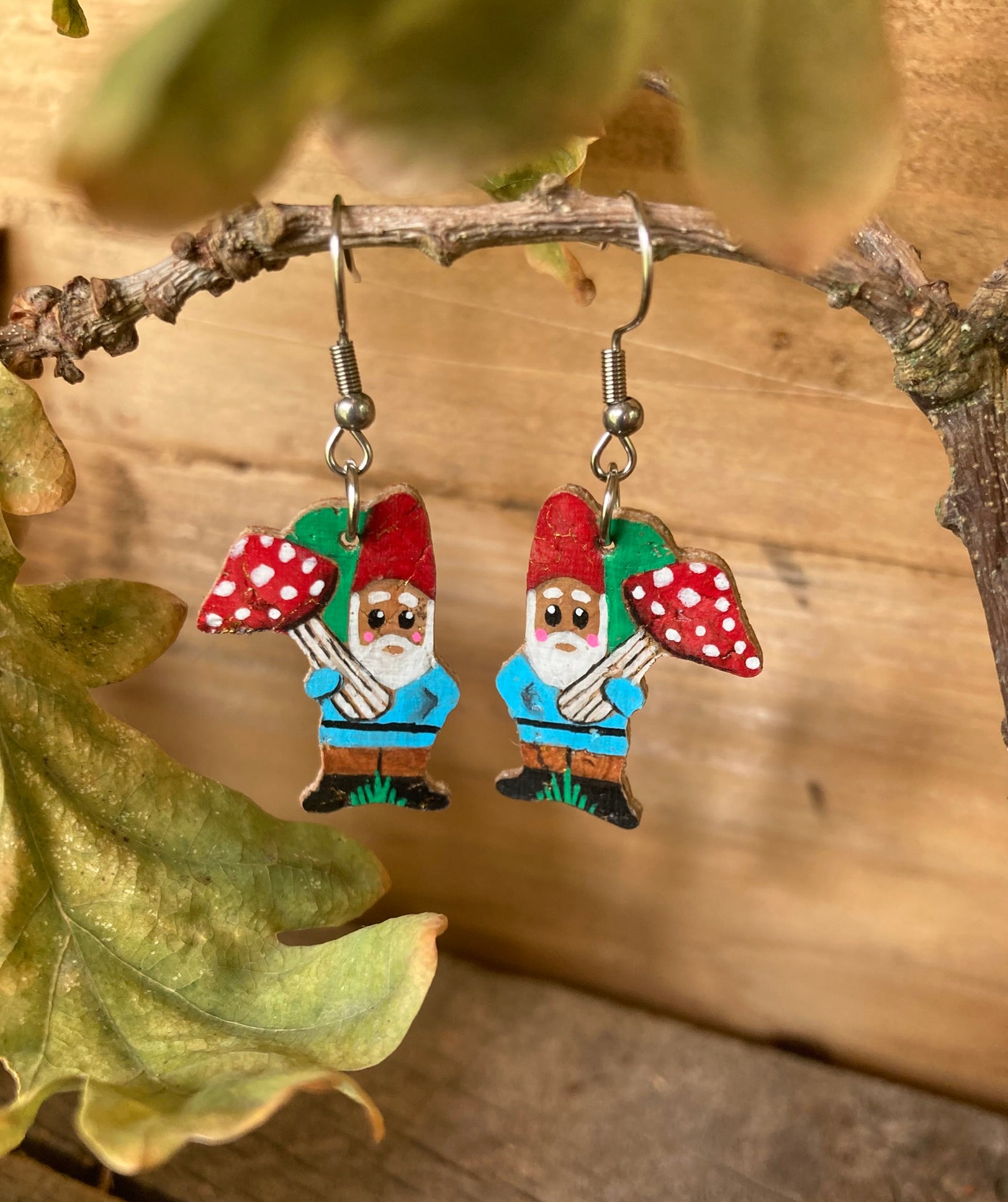 Handpainted gnome and toadstool cork earrings on silver hooks hanging from a woodland branch with green foliage — sustainable cork jewellery