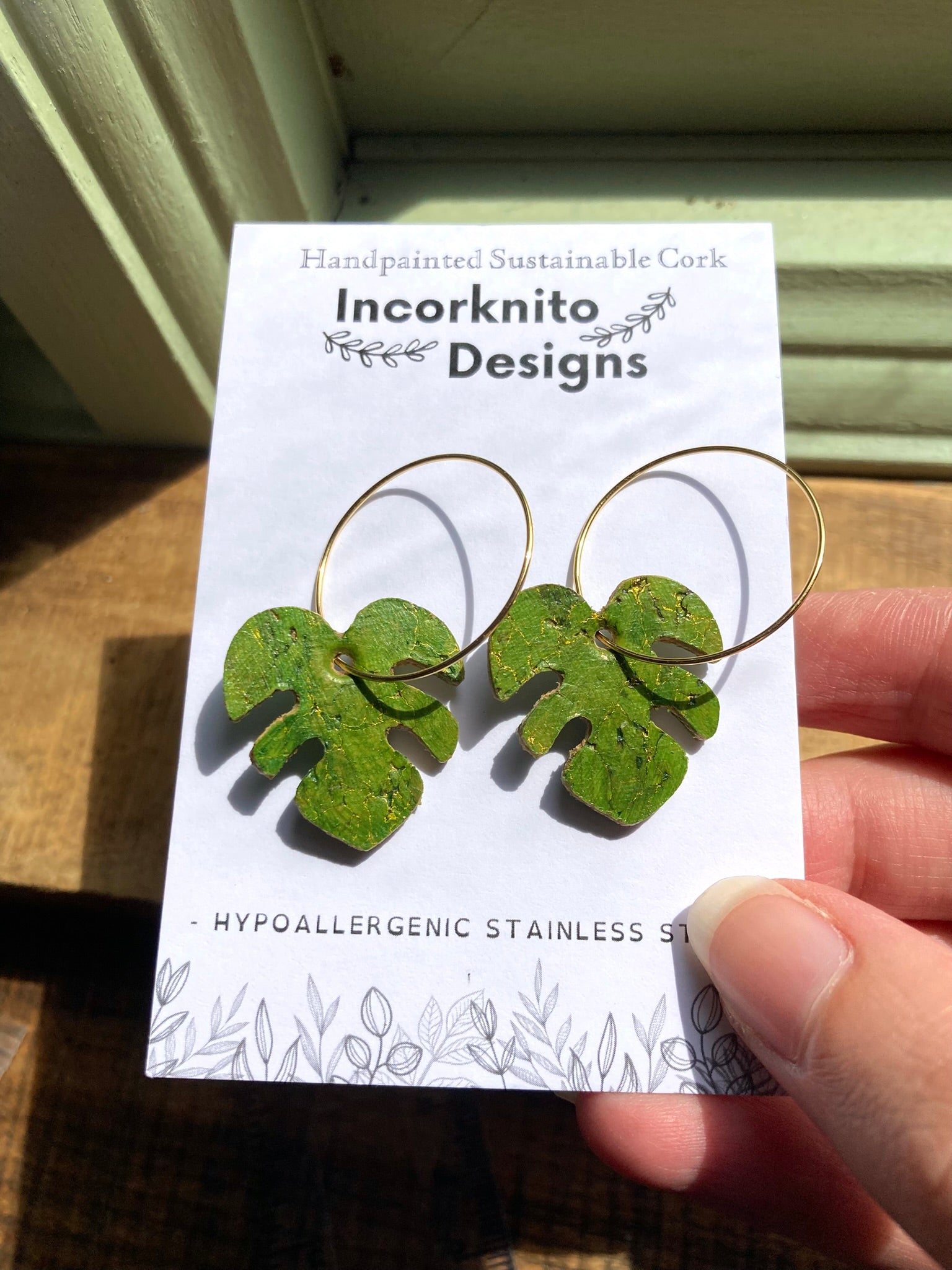 Green monstera cork earrings on gold hoops on Incorknito Designs branded card — handmade sustainable plant jewellery handmade in Suffolk UK