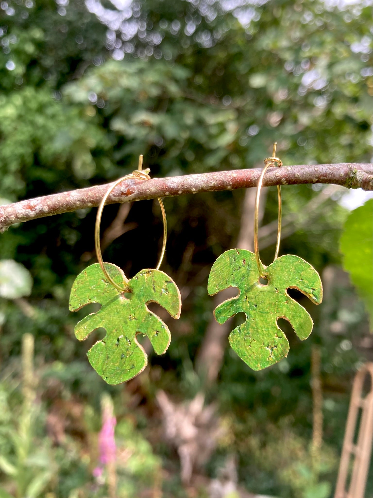 Handpainted green monstera cork earrings on gold hoops hanging from a garden branch — vivid olive green and gold fleck detail catching natural light