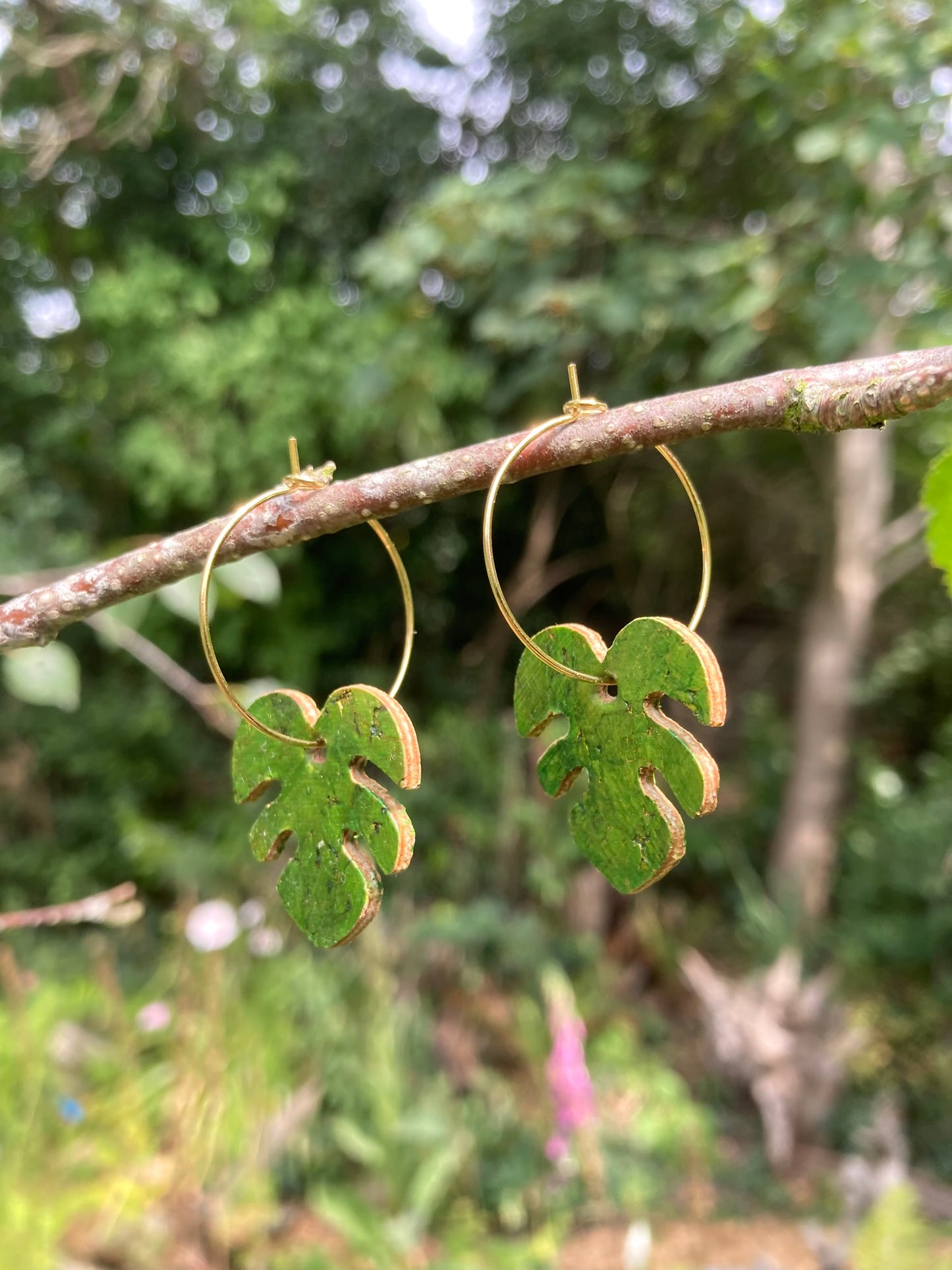 Sideways view of handpainted green monstera cork earrings on gold hoops suspended from a woodland branch — khaki green and gold against a lush garden backdrop