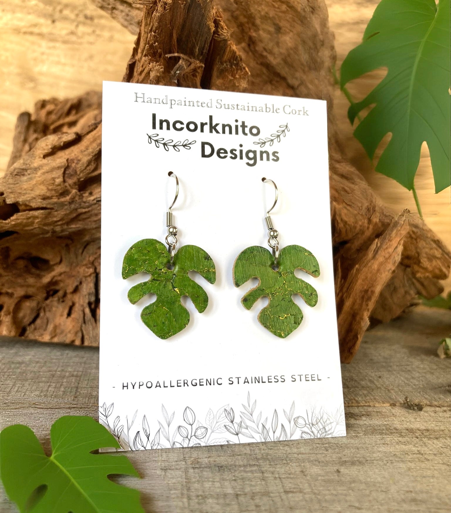 Green monstera cork earrings on silver hooks on Incorknito Designs branded card propped against driftwood with monstera leaves — hypoallergenic sustainable jewellery
