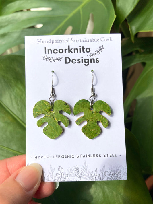 Handpainted green monstera cork earrings on silver hooks on Incorknito Designs branded card, held against real monstera leaves — sustainable plant jewellery handmade in Suffolk UK