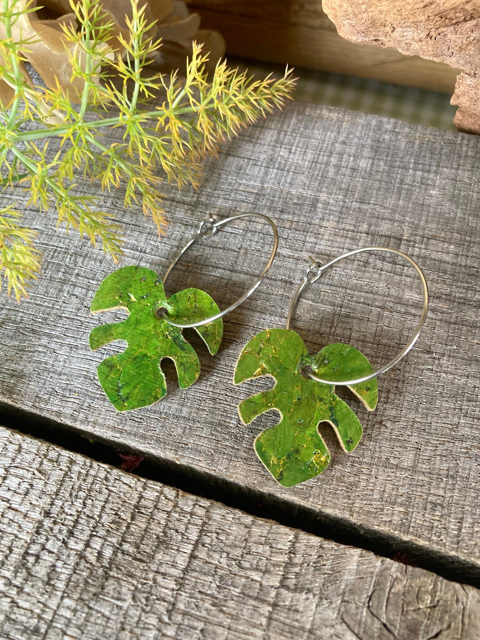 Green monstera cork earrings on silver hoops flat lay on weathered grey wood with yellow-green fennel sprigs — handpainted eco-friendly plant jewellery