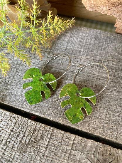 Green monstera cork earrings on silver hoops flat lay on weathered grey wood with yellow-green fennel sprigs — handpainted eco-friendly plant jewellery