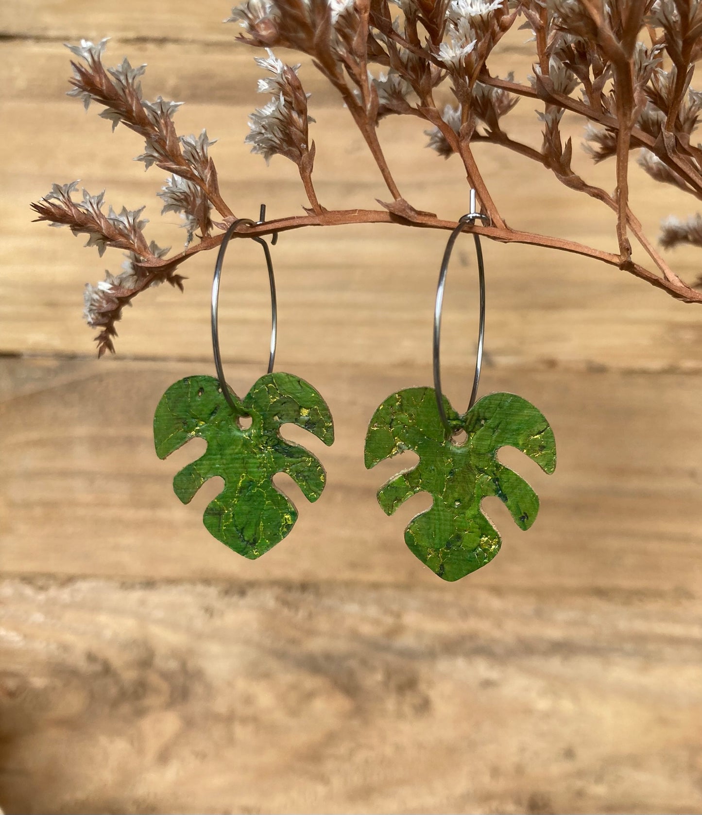 Handpainted green monstera cork earrings on silver hoops hanging from a russet dried botanical branch — gold fleck cork detail catching warm light