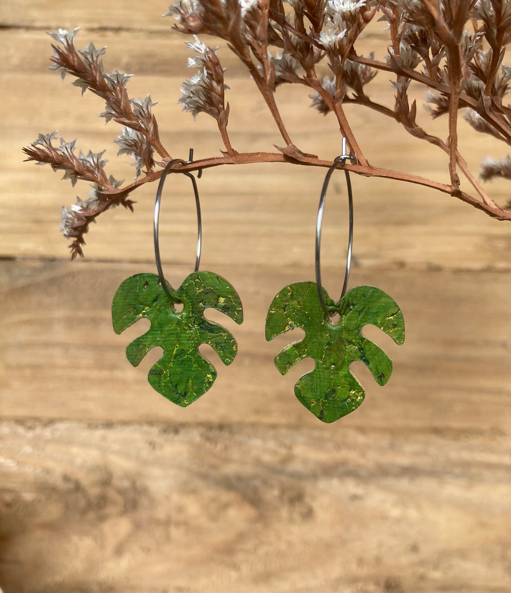 Handpainted green monstera cork earrings on silver hoops hanging from a russet dried botanical branch — gold fleck cork detail catching warm light
