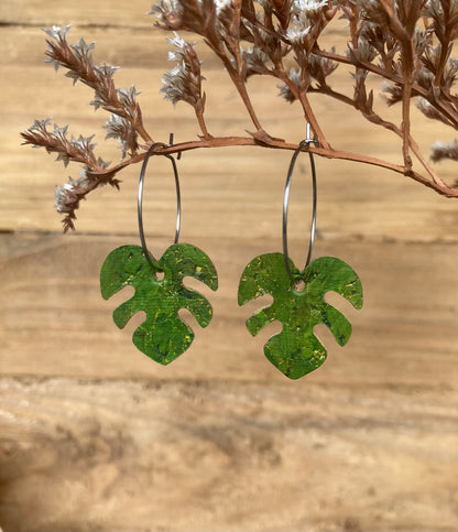 Handpainted green monstera cork earrings on silver hoops hanging from a russet dried botanical branch — gold fleck cork detail catching warm light