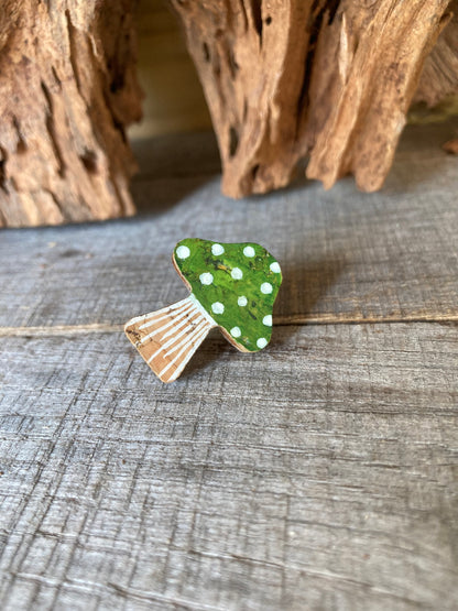 Handpainted green toadstool cork pin badge on weathered grey wood with driftwood behind — rich woodland green spotted cap with gold fleck shimmer, handmade in Suffolk UK