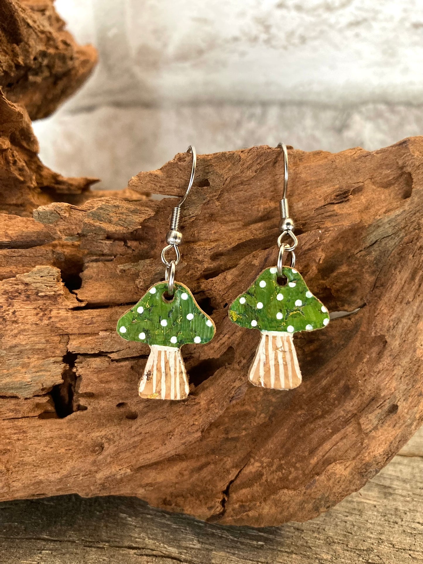 Green toadstool cork medium earrings resting against driftwood — silver hooks showing the natural cork texture and handpainted green cap