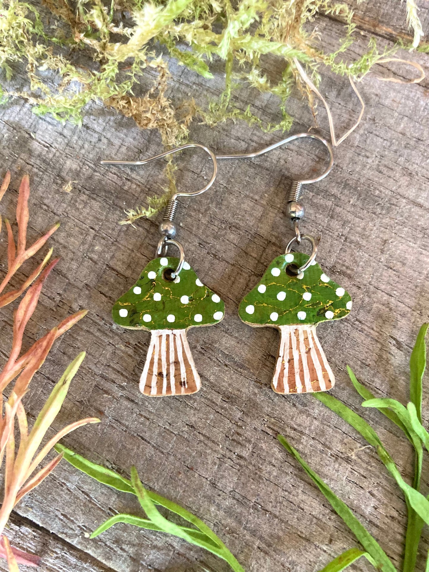 Green toadstool cork medium earrings flat lay on weathered wood with red botanicals — gold fleck cork with handpainted green cap and white polka dots