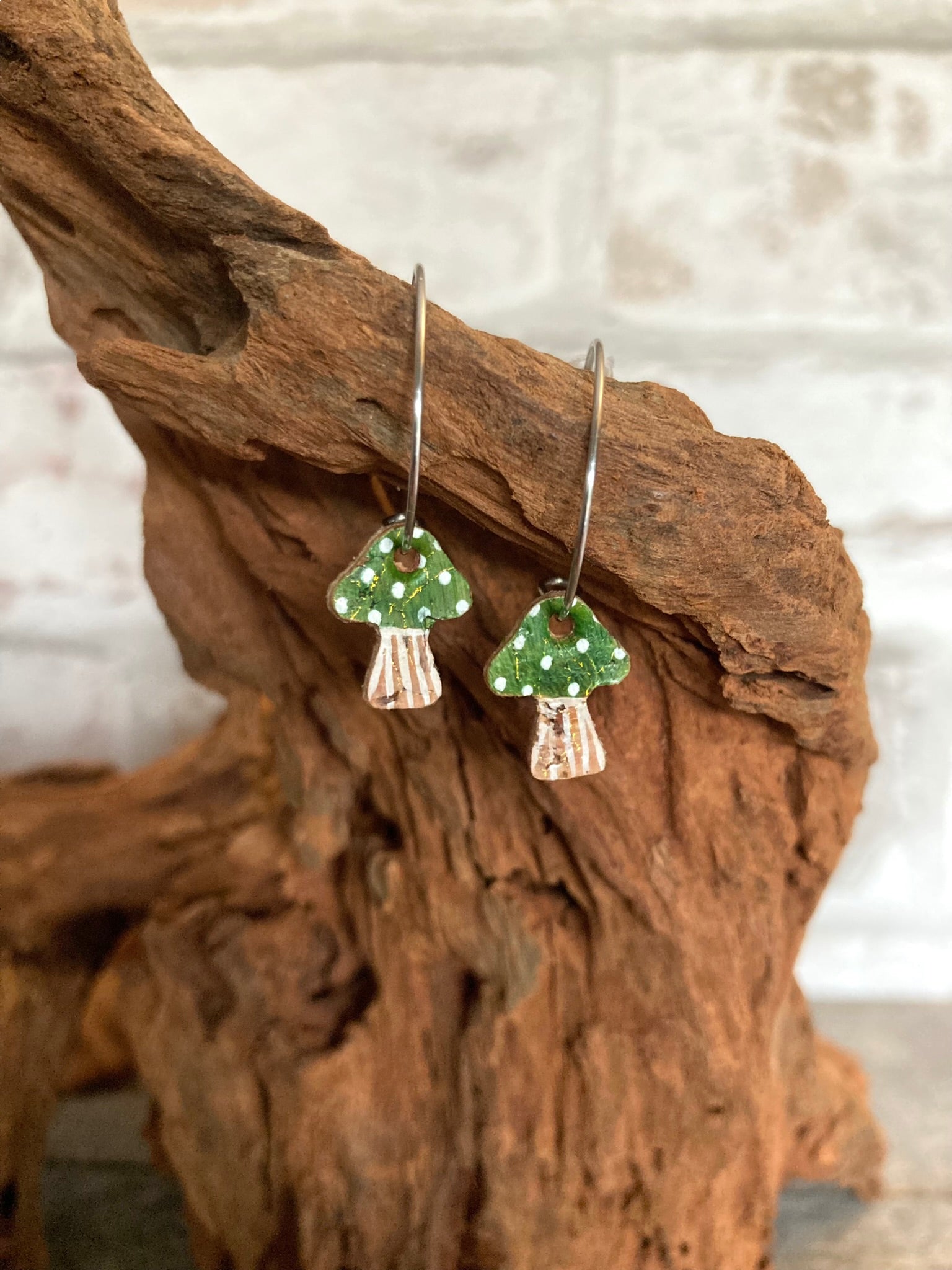 Green toadstool cork mini hoop earrings hanging from driftwood against a whitewashed wall — lightweight woodland jewellery for nature lovers