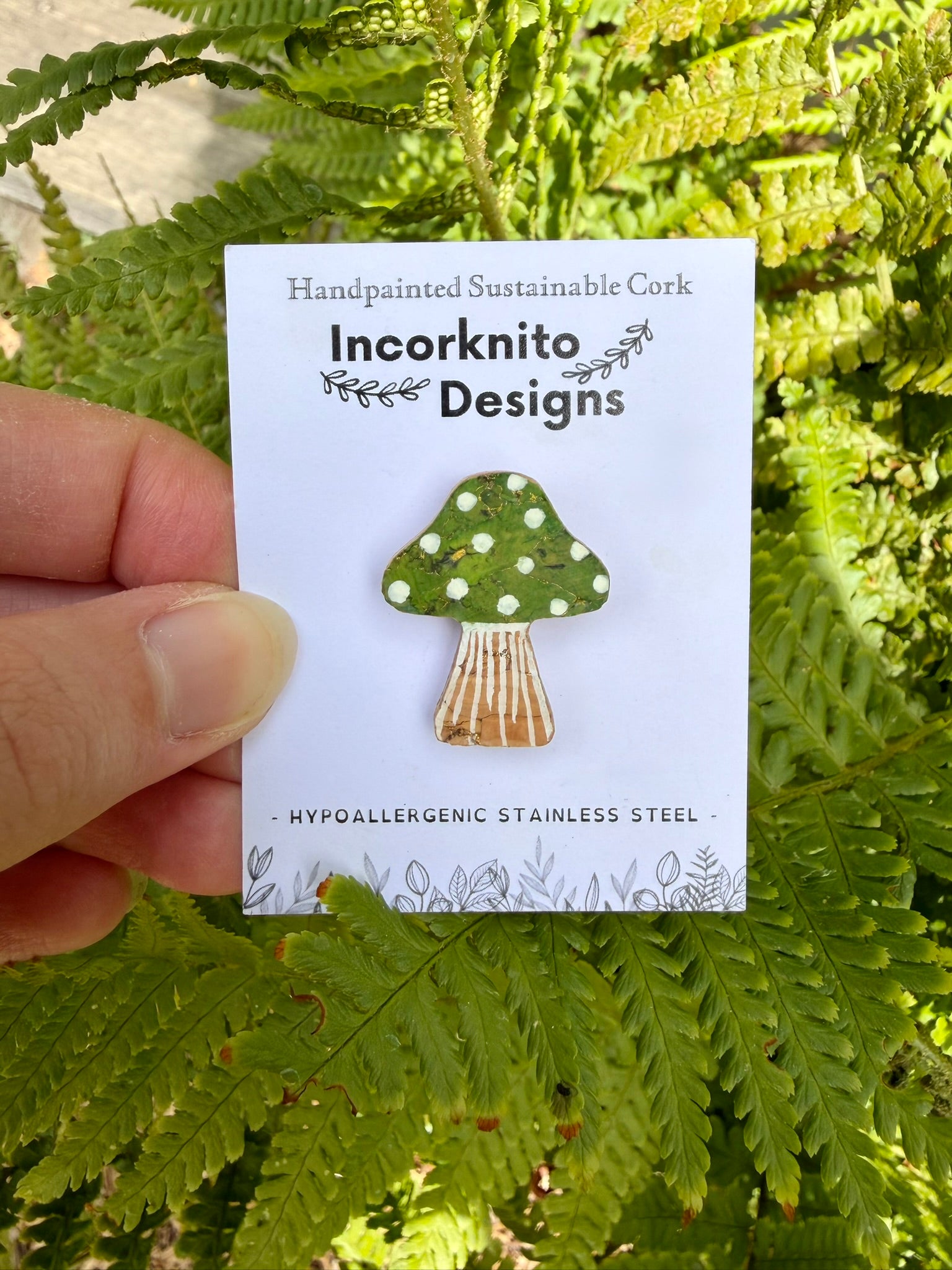 Green toadstool with white polka dots  and gold flecked cork pin badge on Incorknito Designs branded card held against green ferns — handpainted sustainable cork badge handmade in Suffolk UK