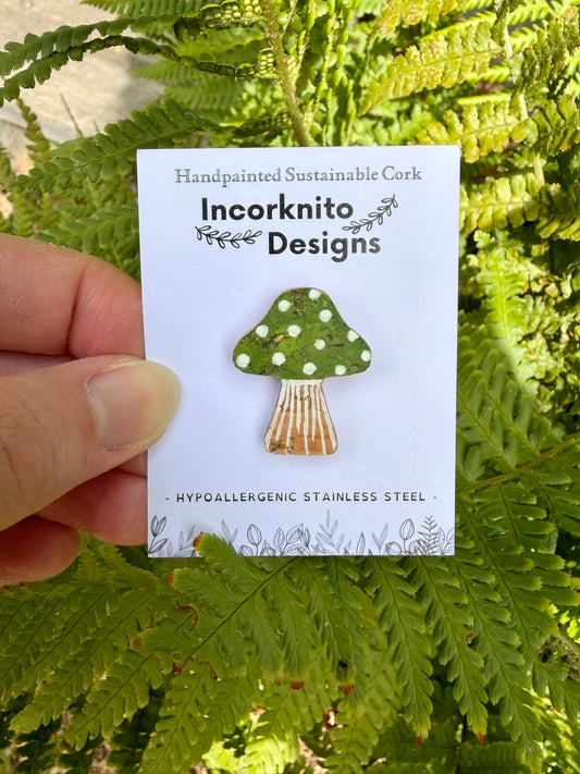 Green toadstool with white polka dots  and gold flecked cork pin badge on Incorknito Designs branded card held against green ferns — handpainted sustainable cork badge handmade in Suffolk UK