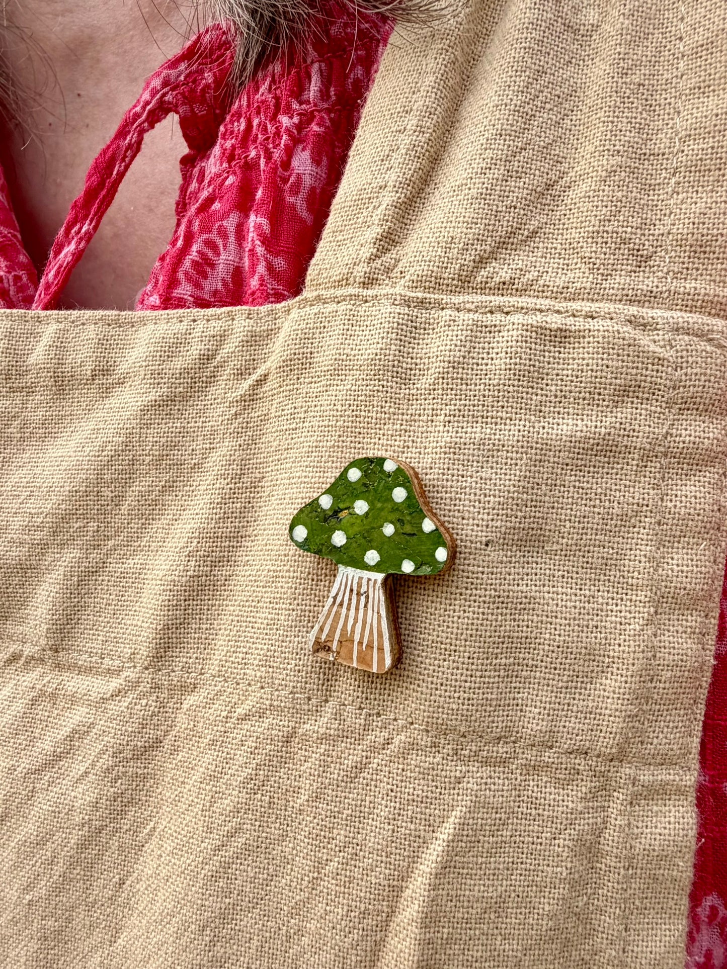 Green toadstool cork pin badge pinned to a linen apron — handpainted green cap with white spots, sustainable cork badge
