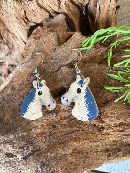 Handpainted grey and white horse cork earrings resting on driftwood — eco-friendly equestrian jewellery handmade in UK