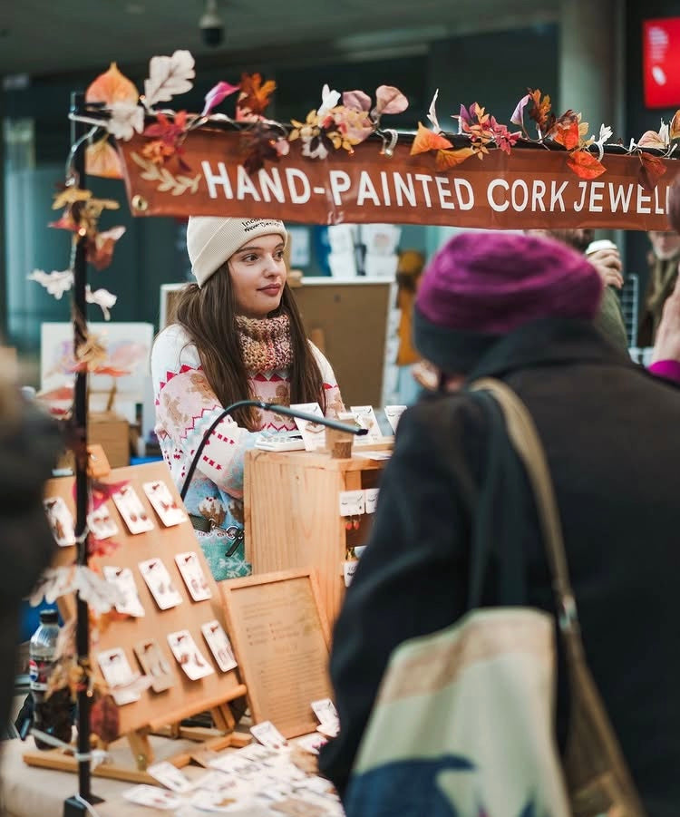 A craft fair booth displaying hand-painted cork jewellery, with a wooden sign reading 'HAND-PAINTED CORK JEWELRY' and autumn-themed decorations. A woman in a white beanie and patterned sweater stands behind the counter, while a person in a dark coat and purple beanie stands admiring products in the foreground.