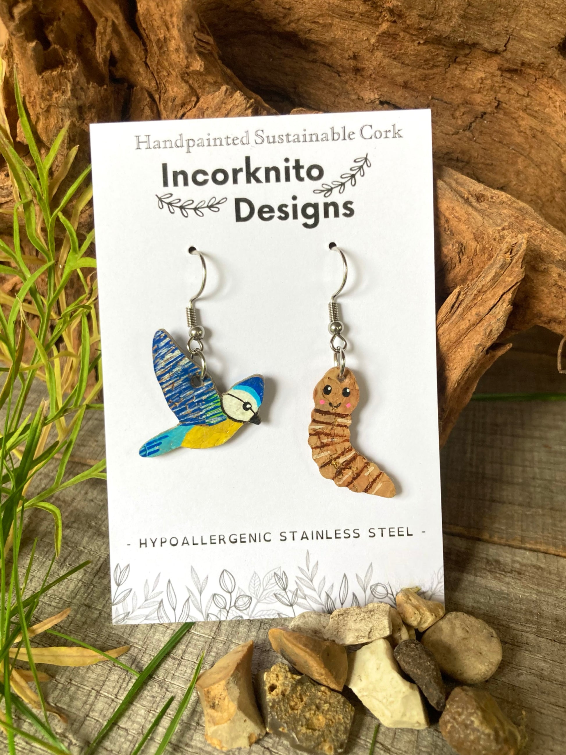 Incorknito Designs blue tit and worm earrings on branded card with green garden foliage, pebbles and wooden background