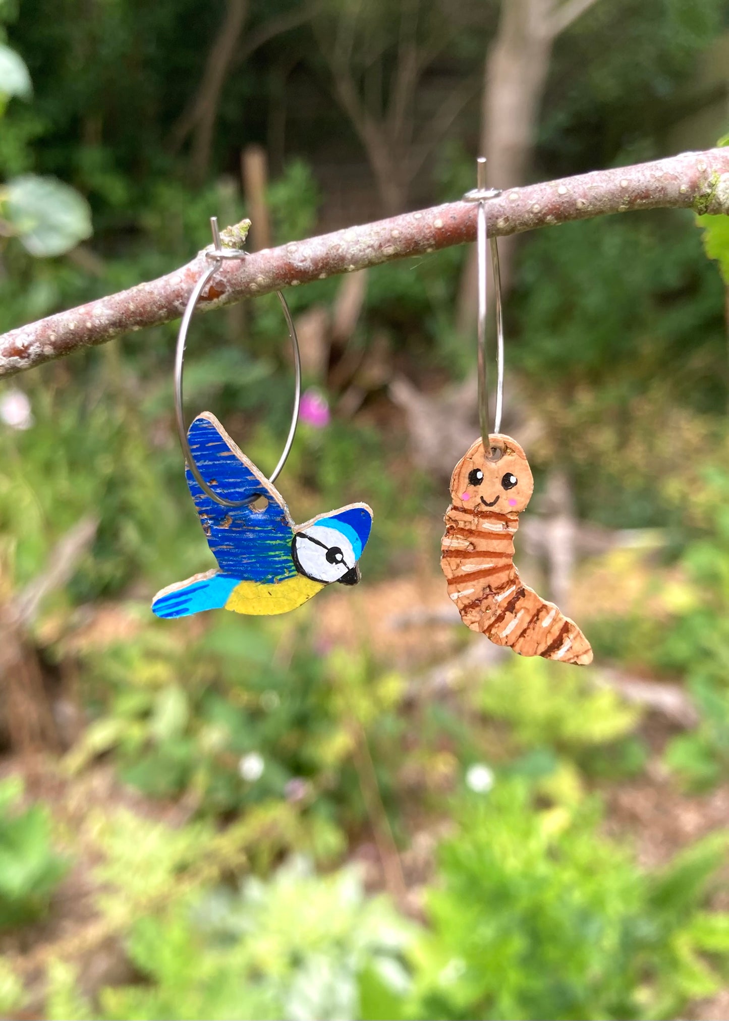 Blue tit and worm mismatched cork earrings hanging from branch on hoop fastenings— early bird catches the worm design. Mimicking a flying bird how it hangs.