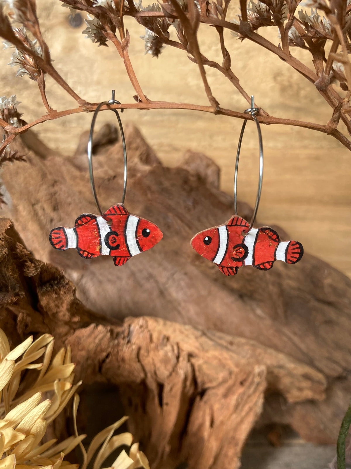 Handpainted clownfish cork earrings on silver hoops, handmade in Suffolk UK — sustainable ocean jewellery hanging from driftwood with dried botanicals