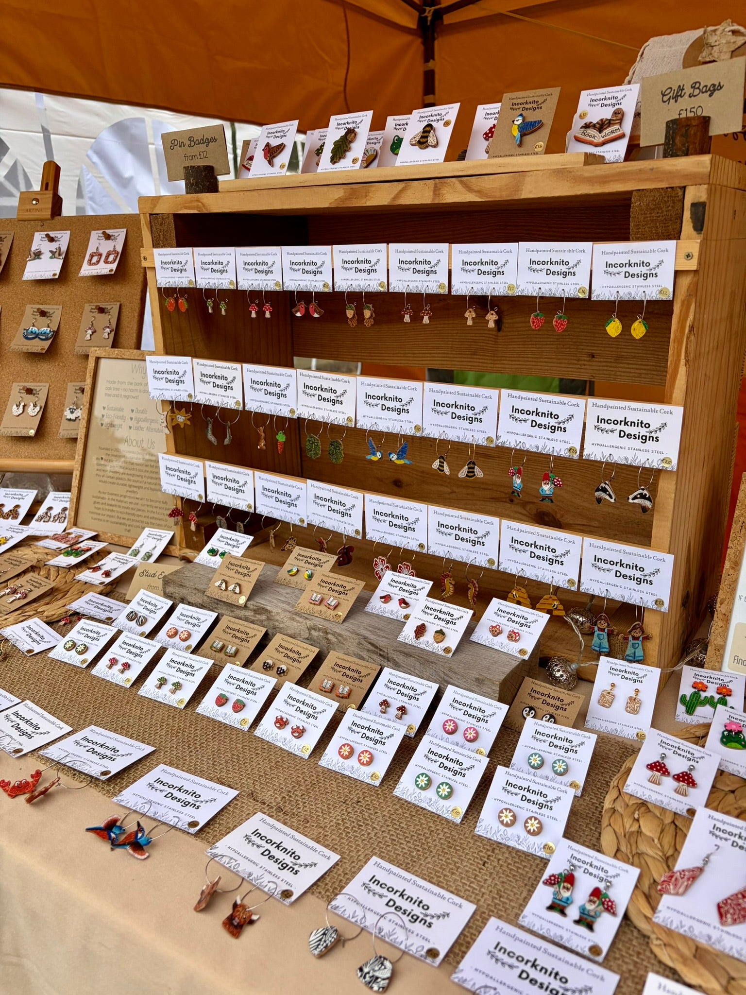 A wooden display stand at an outdoor market showcasing handmade earrings from 'Incorknito Designs' on small white cards, with a tan tent canopy overhead.