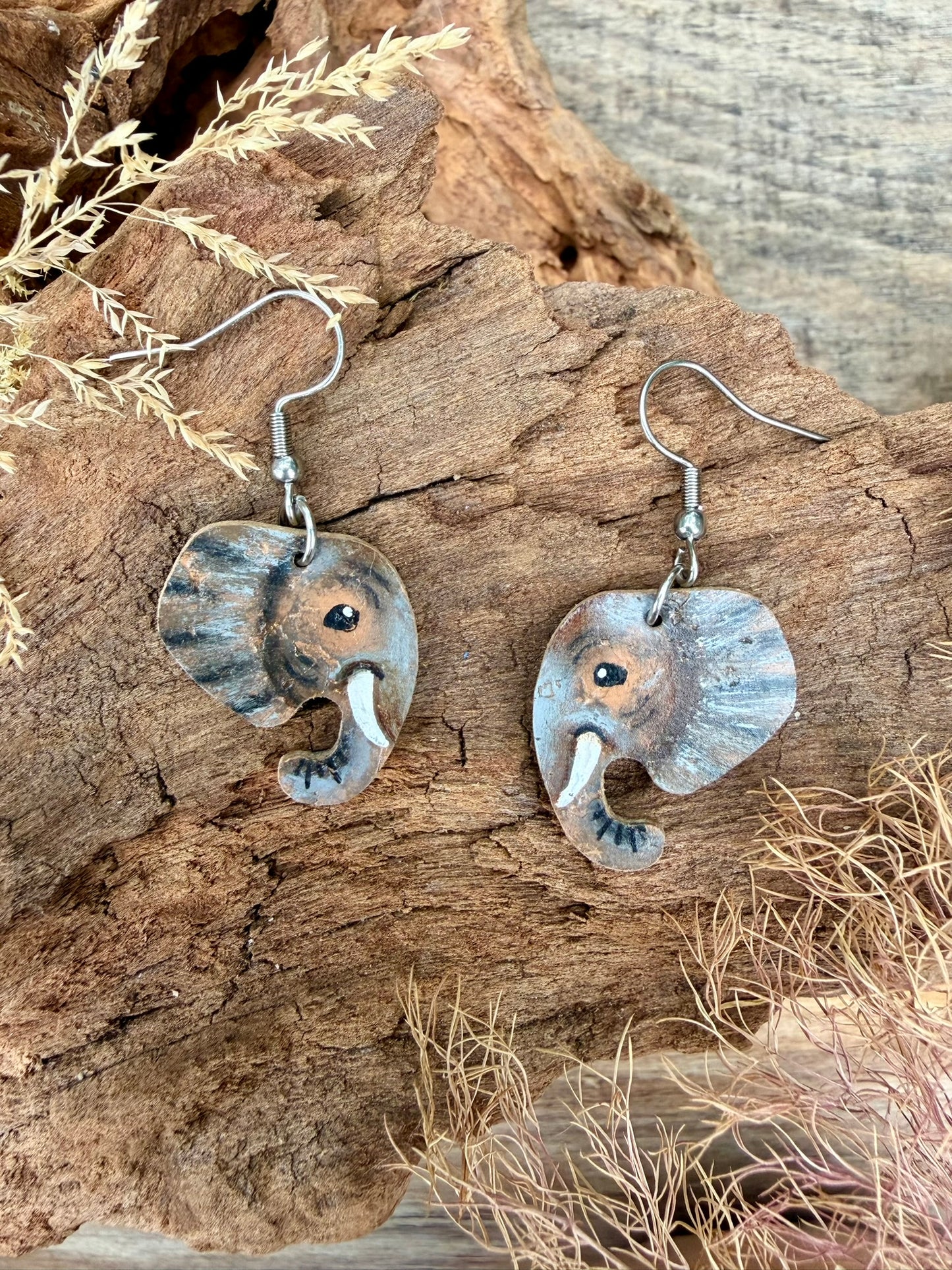 A pair of handmade elephant-shaped drop earrings with a weathered grey and brown finish, featuring silver toned fishhook wires. The earrings are displayed on a piece of natural driftwood with dried grass.