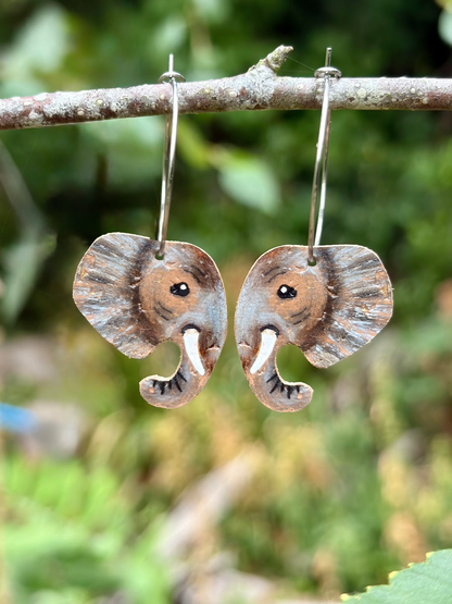 A pair of handmade elephant-shaped dangle earrings hanging from a tree branch outdoors, with a blurred green natural background.