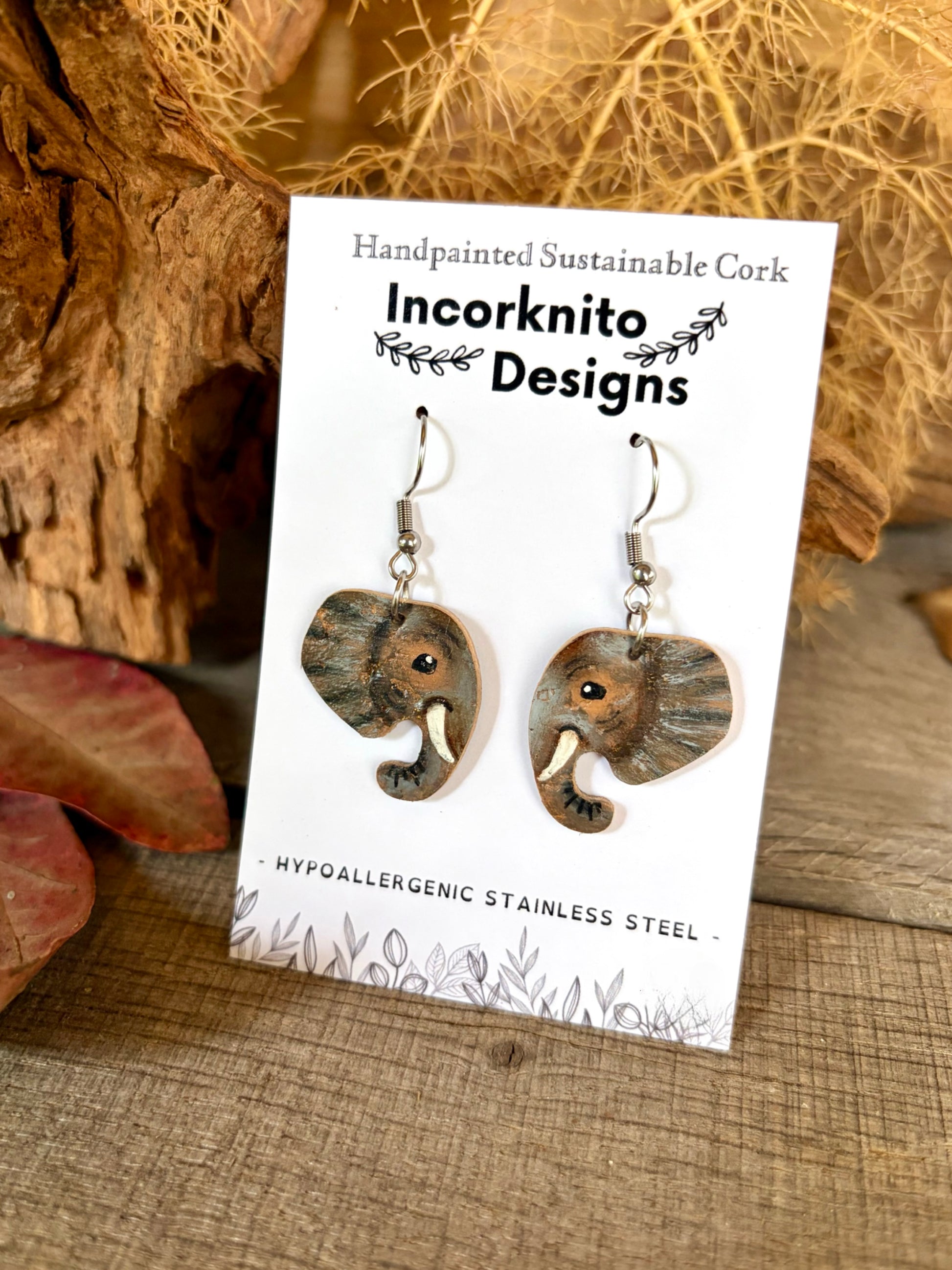 Elephant-shaped dangle earrings displayed on a white card labeled 'Incorknito Designs,' set against a rustic background with wood and dried grass.