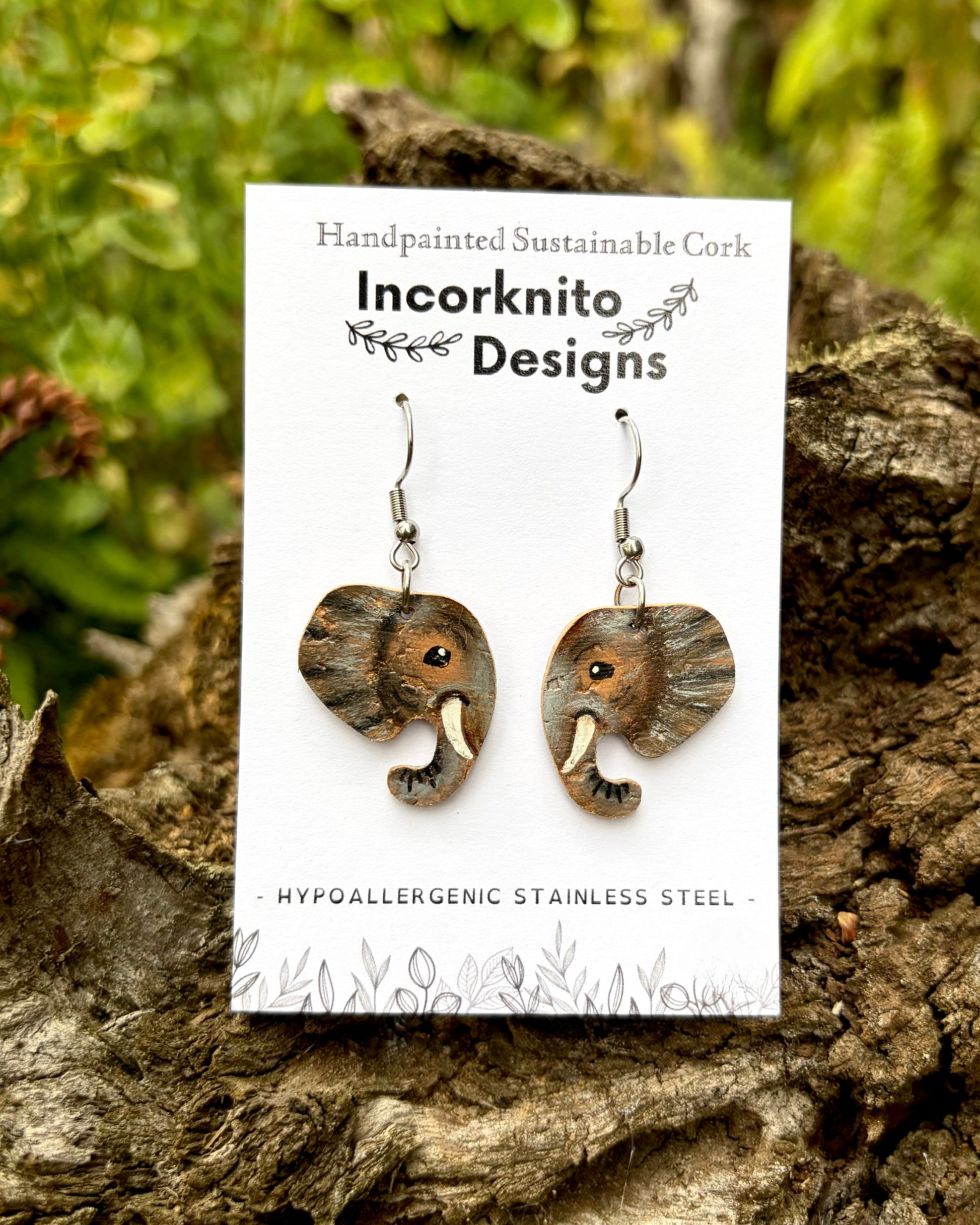 Elephant-shaped dangling earrings made from handpainted sustainable cork and stainless steel, displayed on a white Incorknito Designs card set against a natural outdoor background of tree bark and green foliage.