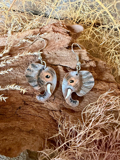 A pair of handmade elephant-shaped dangle earrings with stainless steel hooks, resting on a piece of weathered wood surrounded by dried grass.