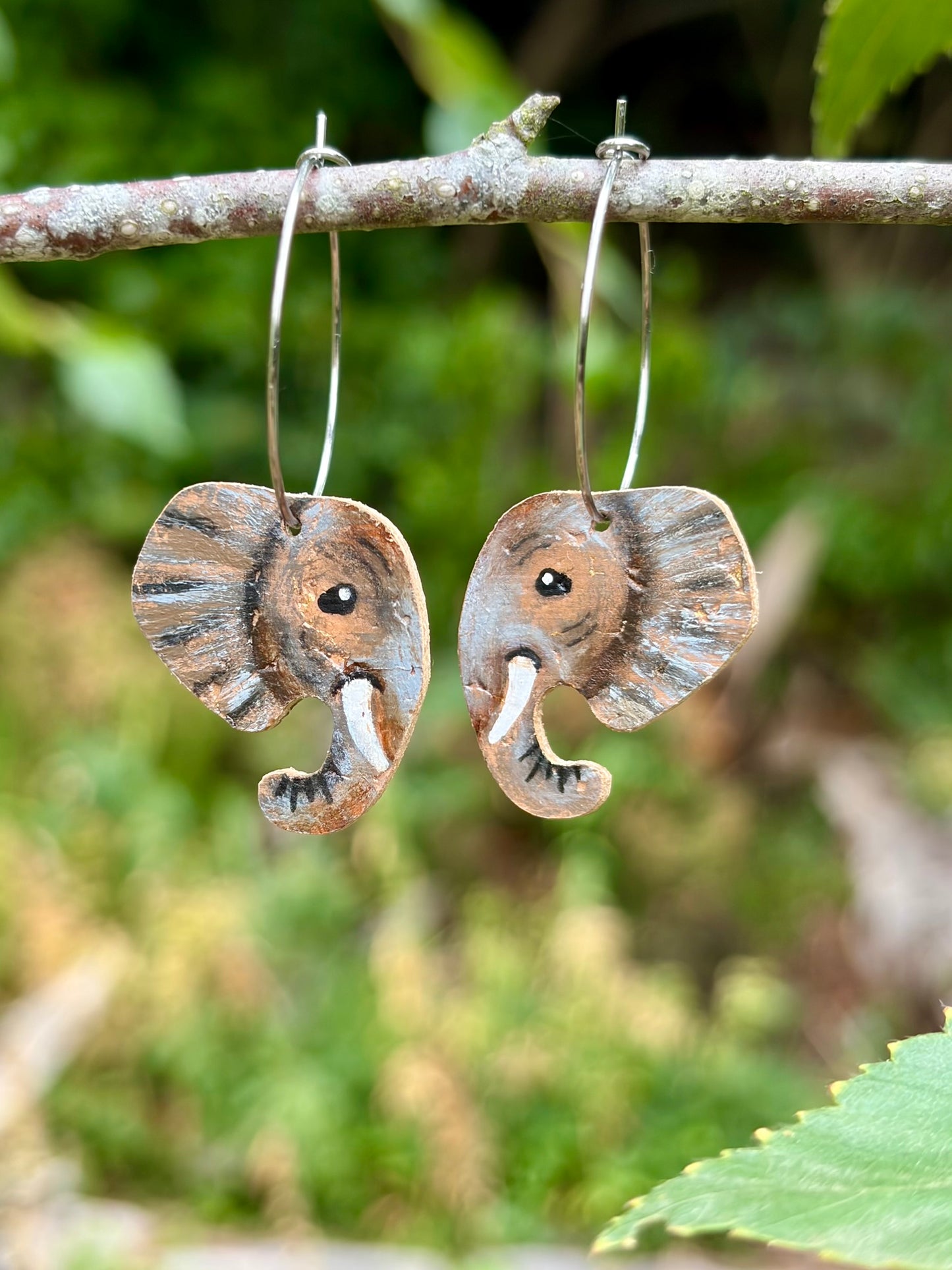 Dangling earrings of elephant shaped heads with textured brown and grey markings and white tusks, hanging from a tree branch outdoors with blurred green foliage in the background.
