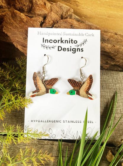 Flying duck earrings on a branded white backing card reading "Incorknito Designs" with a natural wood background and green foliage