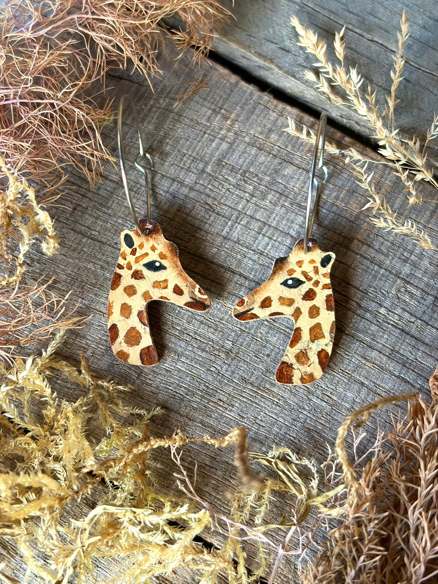 A pair of dangling earrings shaped like giraffe heads, shown on a rustic wooden surface with dried botanicals around them. The giraffes are tan with brown spots, and the hoops are stainless steel