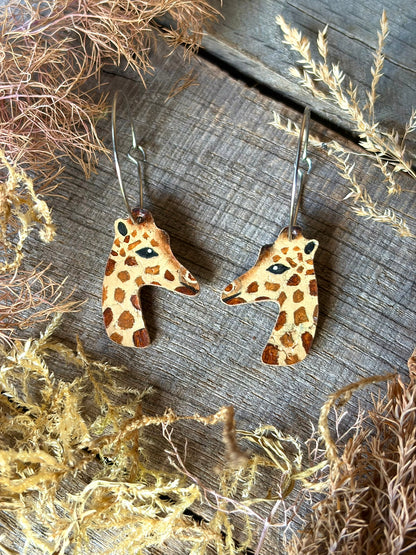 A pair of dangling earrings shaped like giraffe heads, shown on a rustic wooden surface with dried botanicals around them. The giraffes are tan with brown spots, and the hoops are stainless steel