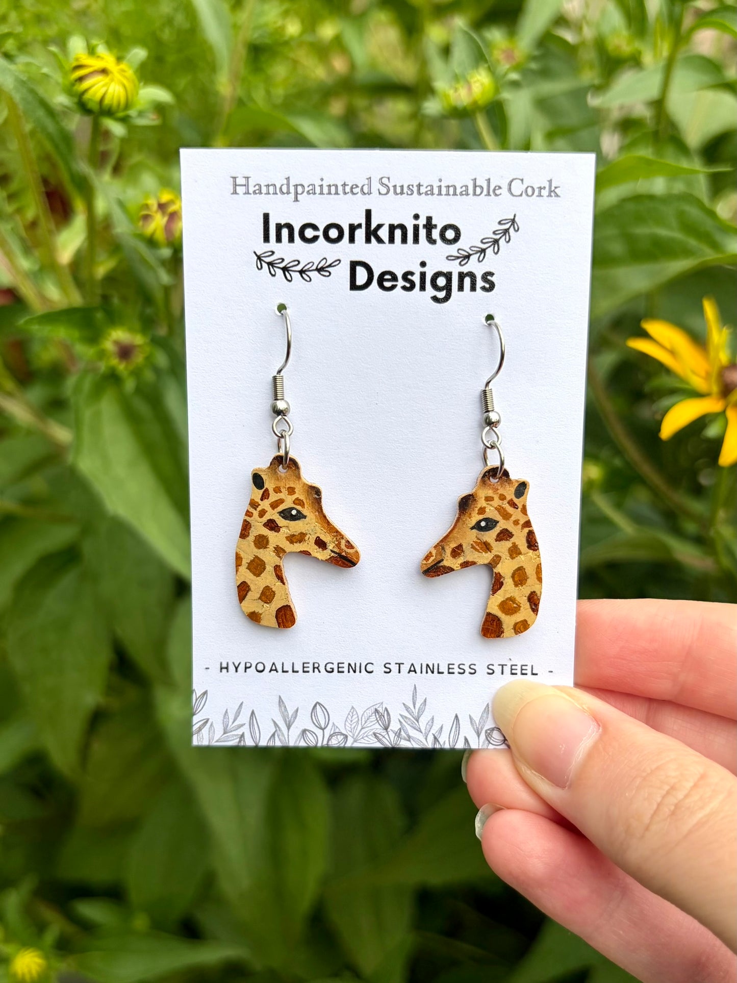 A pair of giraffe-shaped dangling earrings on a white display card held by a hand against a blurred green garden background with yellow flowers. The card reads 'Handpainted Sustainable Cork Incorknito Designs' and 'Hypoallergenic Stainless Steel'.
