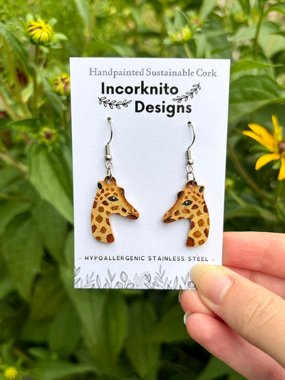 A pair of giraffe-shaped dangling earrings on a white display card held by a hand against a blurred green garden background with yellow flowers. The card reads 'Handpainted Sustainable Cork Incorknito Designs' and 'Hypoallergenic Stainless Steel'.