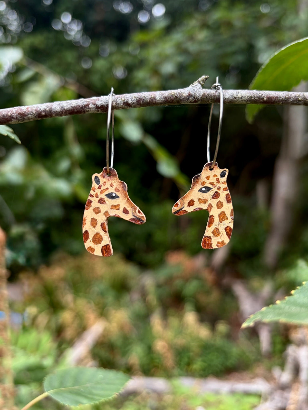 Giraffe cork earrings on silver hoops hanging from a woodland branch against blurred green foliage