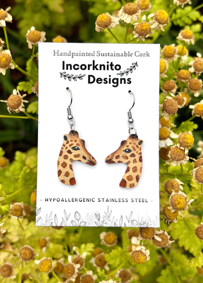 Handcrafted cork giraffe-shaped dangle earrings displayed on a white brand card reading 'Incorknito Designs,' set against a background of green leaves and yellow chamomile flowers.