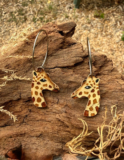 A pair of giraffe-head dangle earrings with stainless steel hoops, displayed on a piece of weathered wood outdoors.