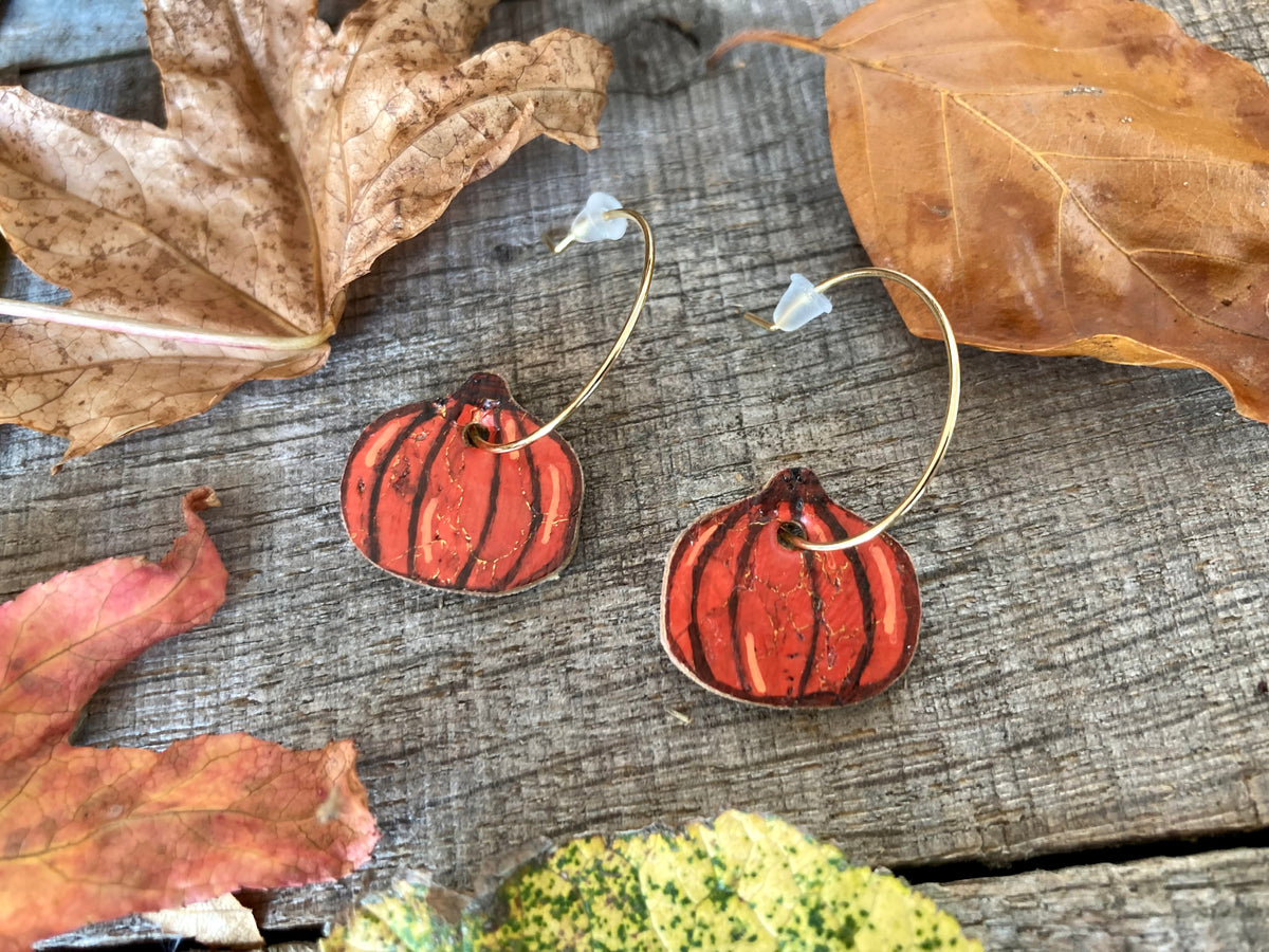 Gold mini hoop pumpkin earrings framed by fallen leaves, showing dark curved segment lines, lighter orange highlights and gold-flecked cork