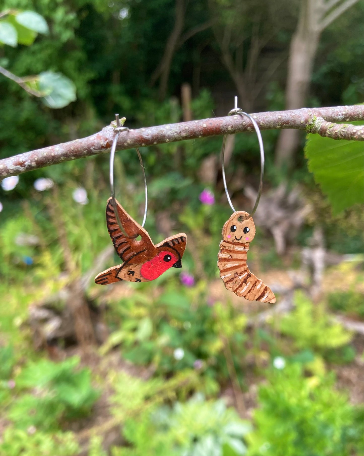 Handpainted cork robin and worm hoop earrings hanging from branch with natural green background