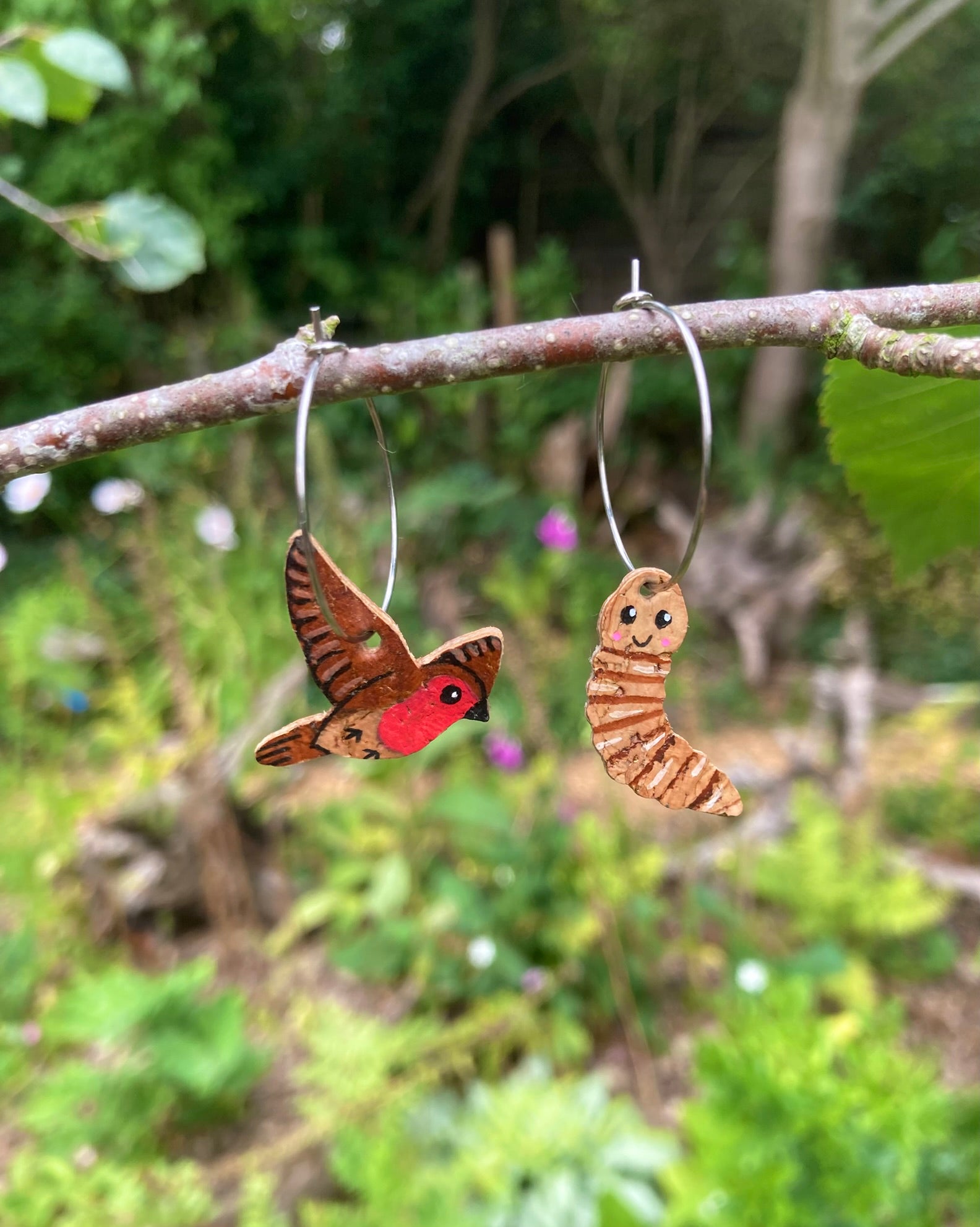 Handpainted cork robin and worm hoop earrings hanging from branch with natural green background