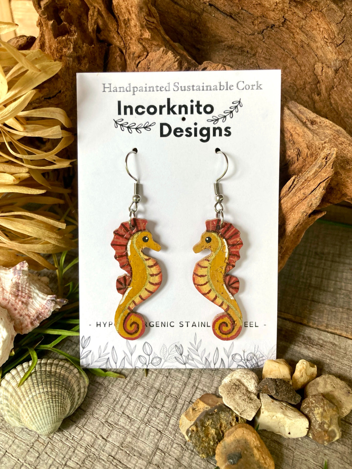 Handpainted sustainable cork seahorse earrings on a white card with 'Incorknito Designs' branding, surrounded by natural elements, including wood, shells and pebbles.