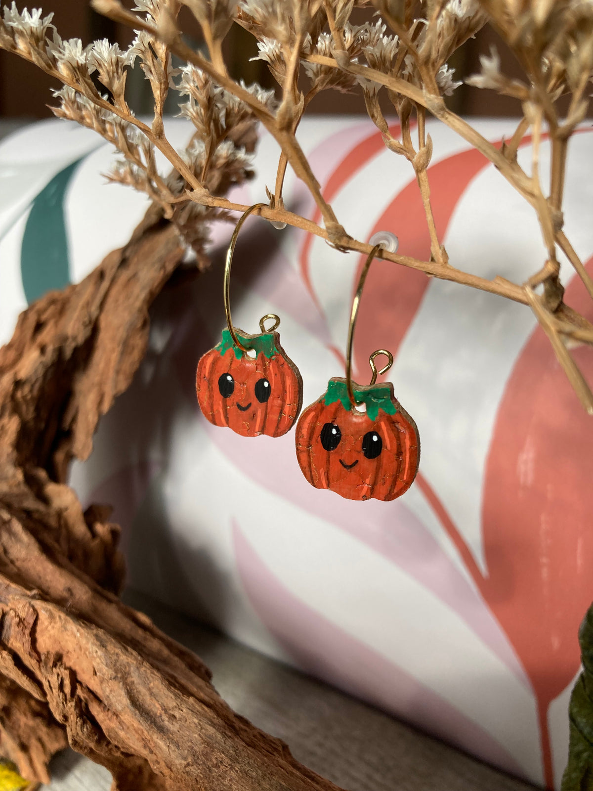 Handpainted pumpkin earrings suspended from a dried flower stem on gold open back hoops, with bark and a patterned cushion behind.