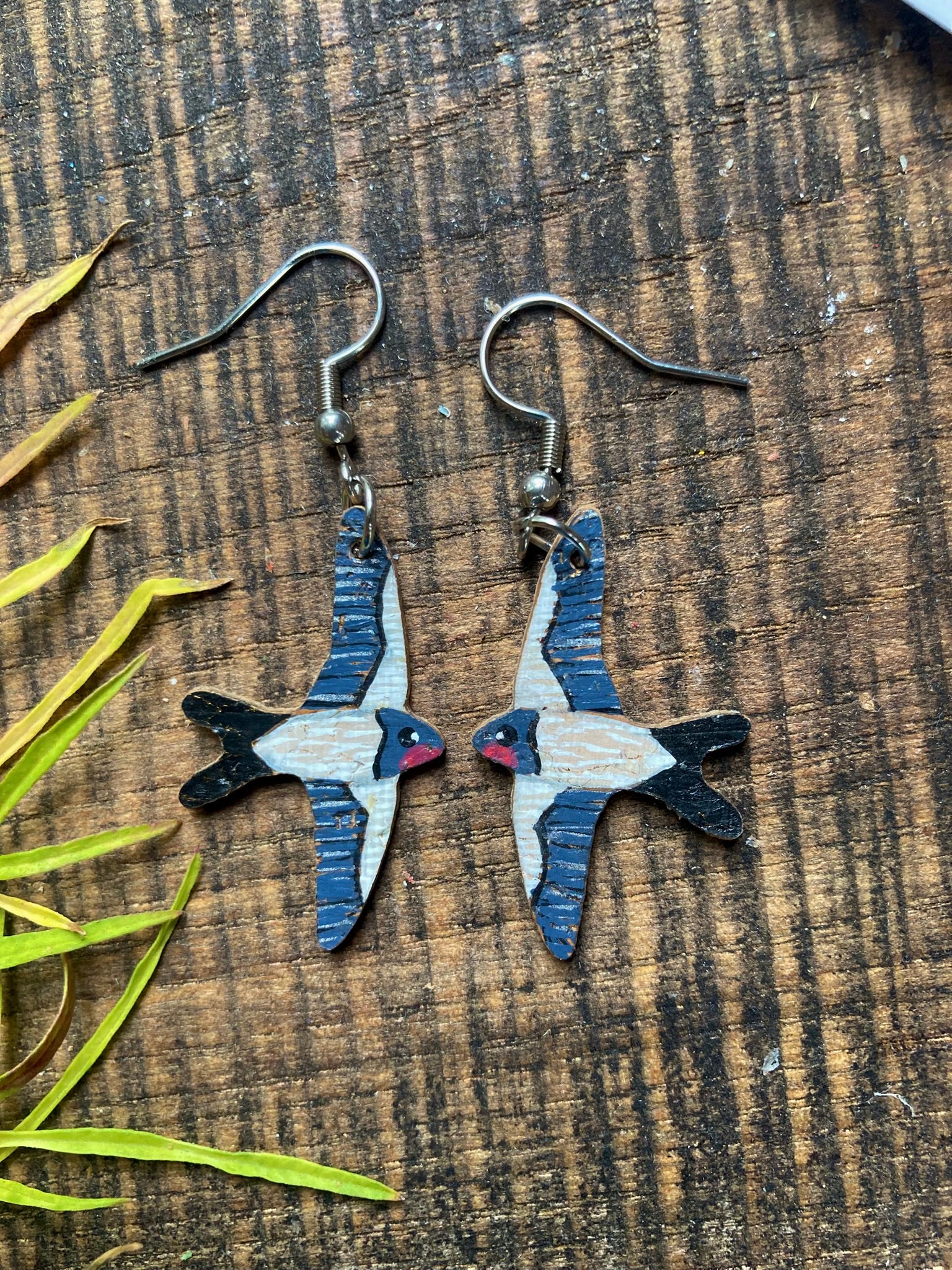 Handpainted swallow earrings on silver hooks on dark wooden surface with yellow leaves